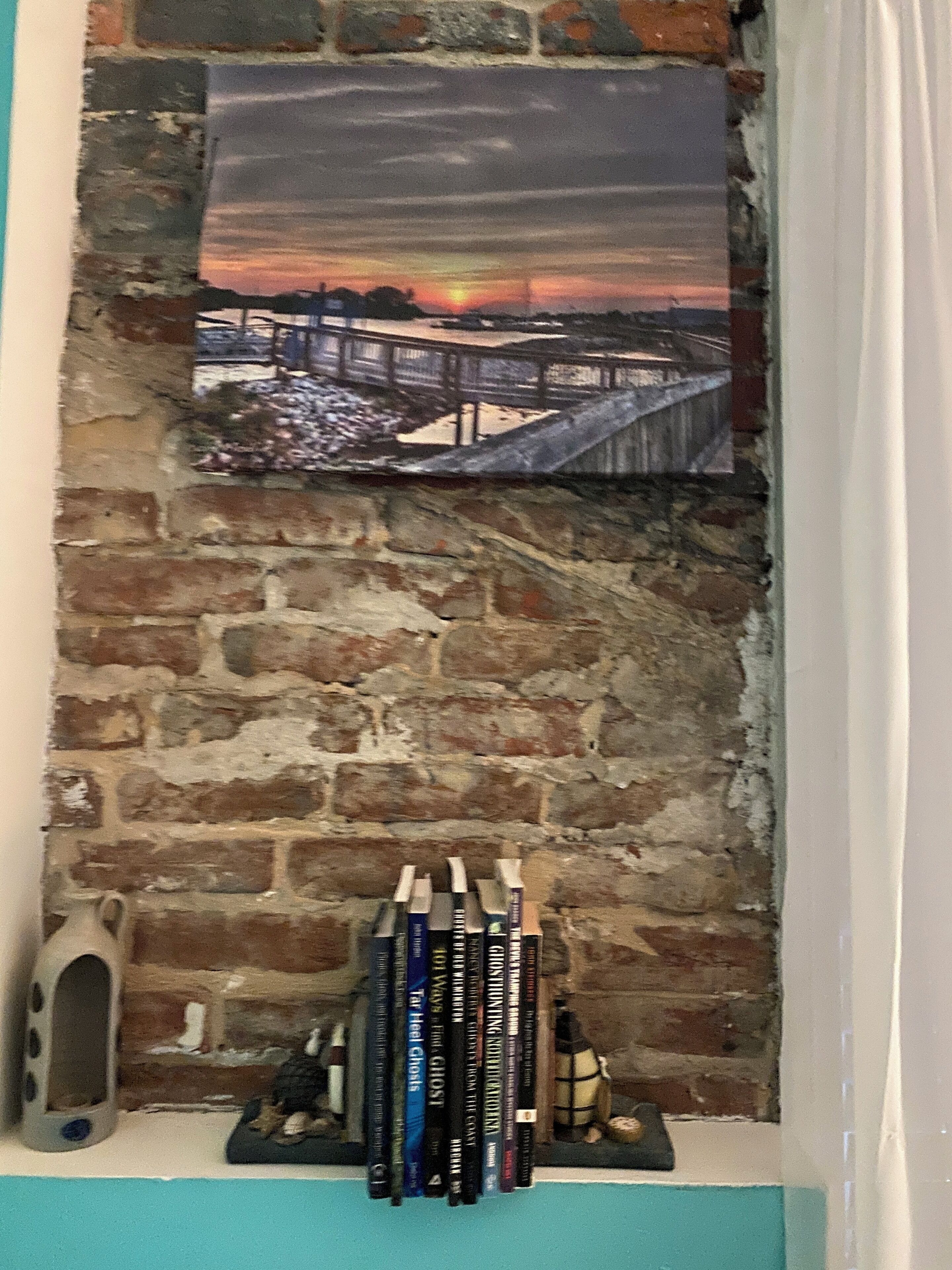 A bookshelf against a brick wall with a sunset painting above it.