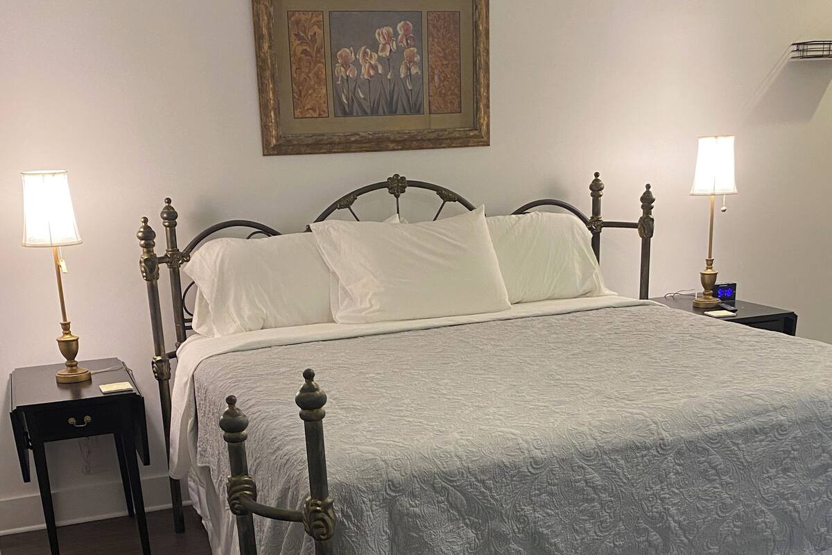 A neatly made bed with a white comforter and pillows, flanked by two bedside tables with lamps, a framed picture on the wall, and a wooden floor.