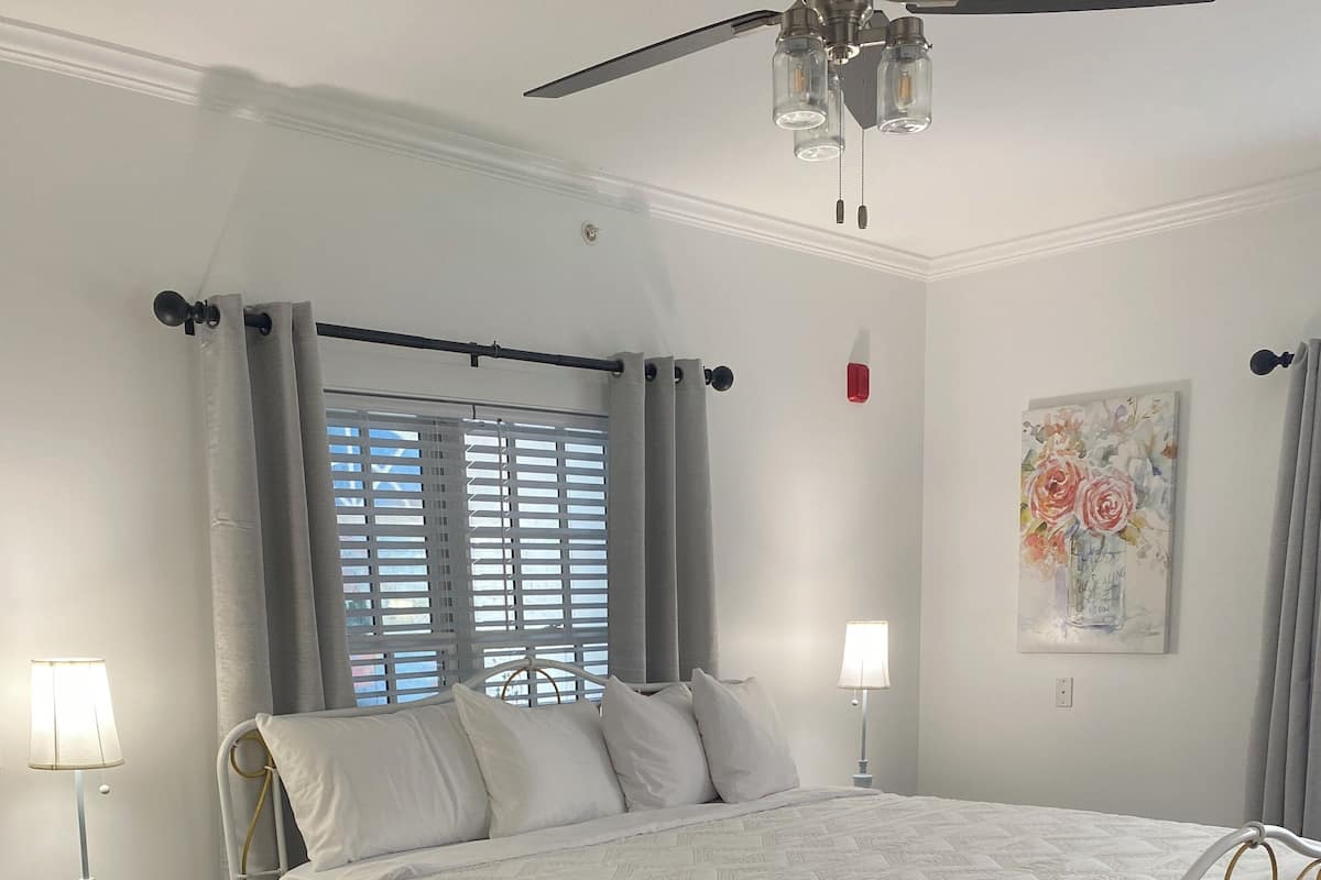 A bedroom with a bed, bedside tables, a ceiling fan, a window with blinds, and a painting on the wall.