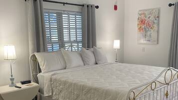 A bedroom with a bed, bedside tables, a ceiling fan, a window with blinds, and a painting on the wall.