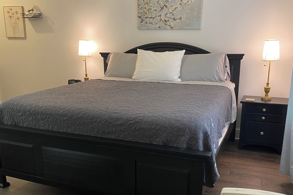 A bedroom with a large bed, two bedside tables with lamps, a nightstand, a chair, a rug, and a wall-mounted painting.