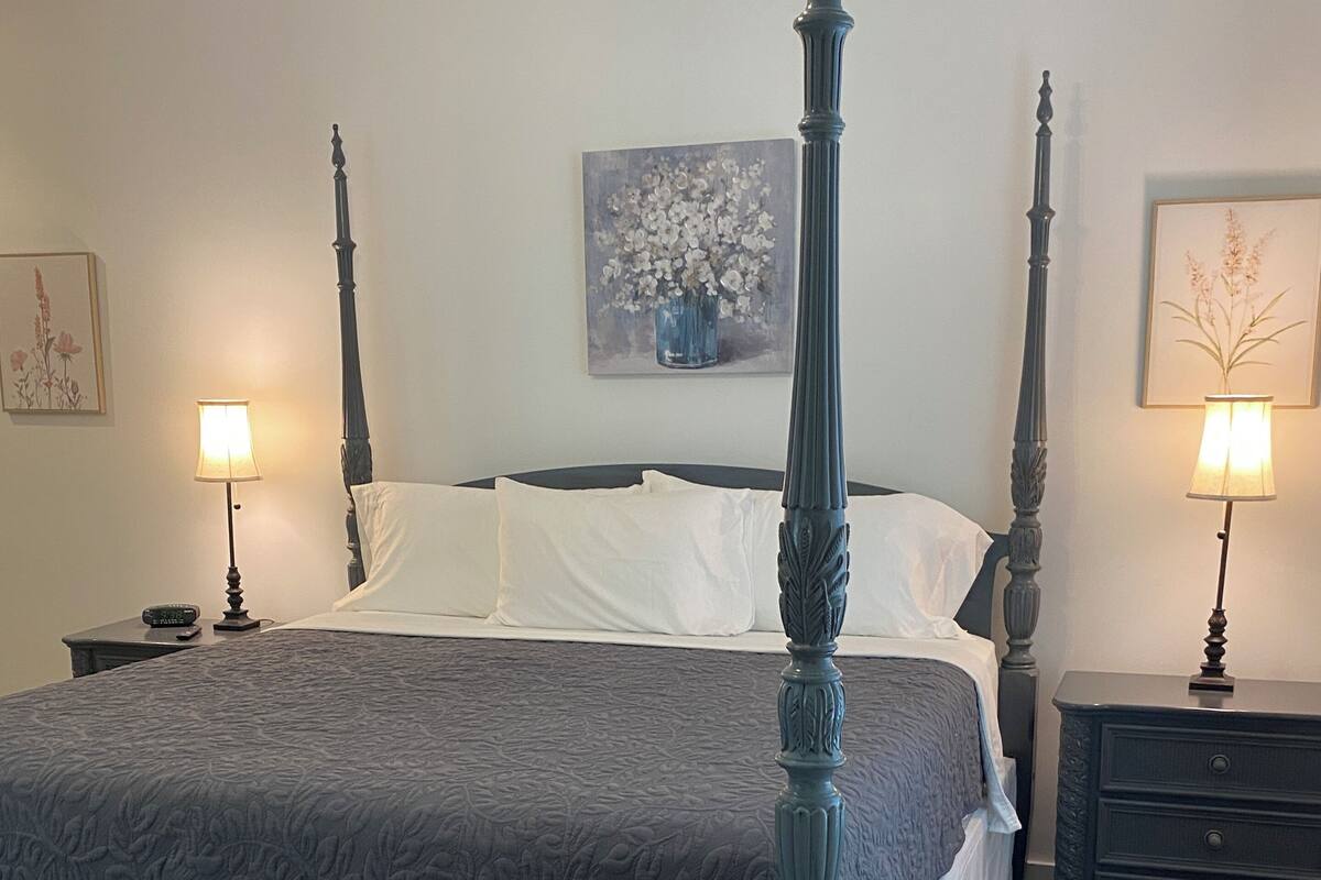 A four-poster bed with a dark headboard and footboard, flanked by two nightstands with lamps. A white upholstered sofa is against the wall, and a framed floral painting hangs above it.