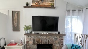 Un soggiorno con camino in pietra, una televisione a schermo piatto montata sopra, un tavolino bianco con oggetti decorativi, un divano grigio con un cuscino decorativo a fantasia e una finestra con tende leggere.