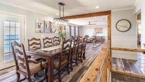 Un espace repas avec une longue table en bois et des chaises, un lustre, et une vue sur l’extérieur grâce à une grande fenêtre.