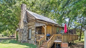 Une cabane rustique en rondins de bois, dotée d’une cheminée en pierre, d’une terrasse en bois et d’un espace salon extérieur.
