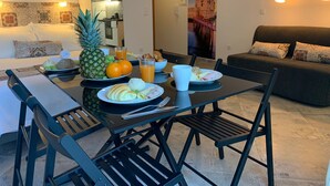 Un coin repas avec une table mise pour le petit-déjeuner, comprenant des assiettes de nourriture, un ananas et des verres de jus d’orange. On aperçoit un canapé et un divan au fond, ainsi qu’une affiche accrochée au mur.