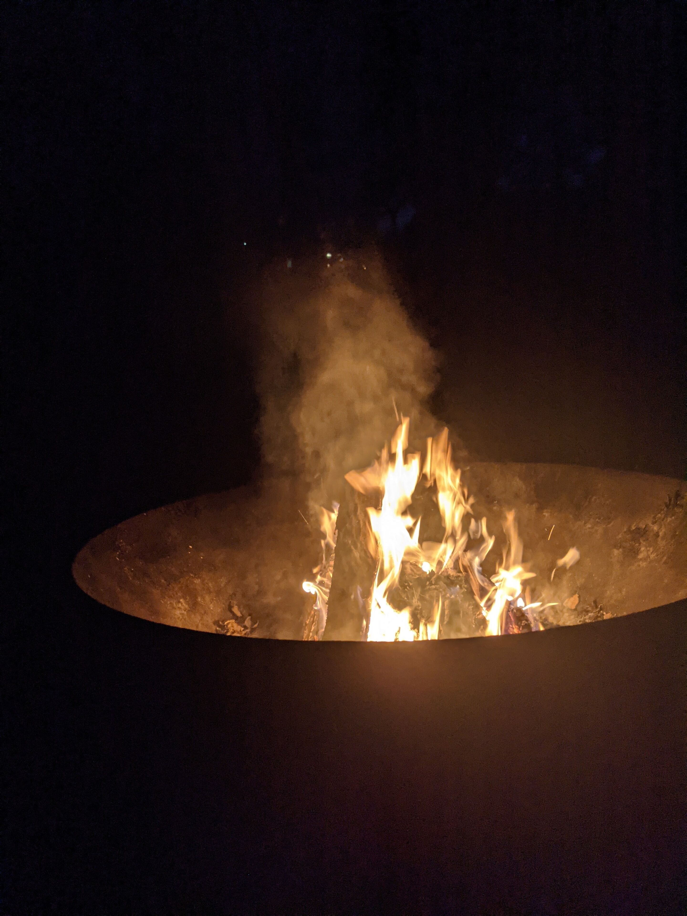 Un feu de camp avec des flammes et de la fumée visibles, entouré d’un foyer circulaire.