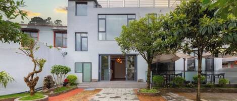 A modern two-story building with a flat roof, large windows, and a paved courtyard with trees and outdoor seating.