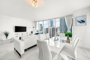 A modern living room with a white sofa, a dining table with chairs, a flat-screen TV, and a city view through large windows. - ICON W BRICKELL MAGNIFICENT RIVER VIEW 2BR/2BA #2102 (Miami)