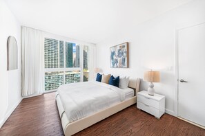 A modern bedroom with a large bed, white nightstands, a lamp, and a city view through floor-to-ceiling windows. - ICON W BRICKELL MAGNIFICENT RIVER VIEW 2BR/2BA #2102 (Miami)