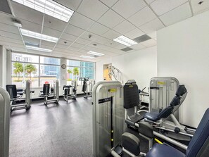 A gym with various exercise machines, large windows with a view of palm trees and buildings, and a clock on the wall. - Brickell 1BR • City Views • Balcony • FREE Parking • Pool • Hot Tub •Gym (Miami)