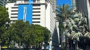 Um edifício alto com uma faixa azul vertical marcante, cercado por palmeiras e outros edifícios.