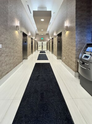 Een moderne hotelgang met liften, een zwart tapijt en een Samsung selfservice kiosk.