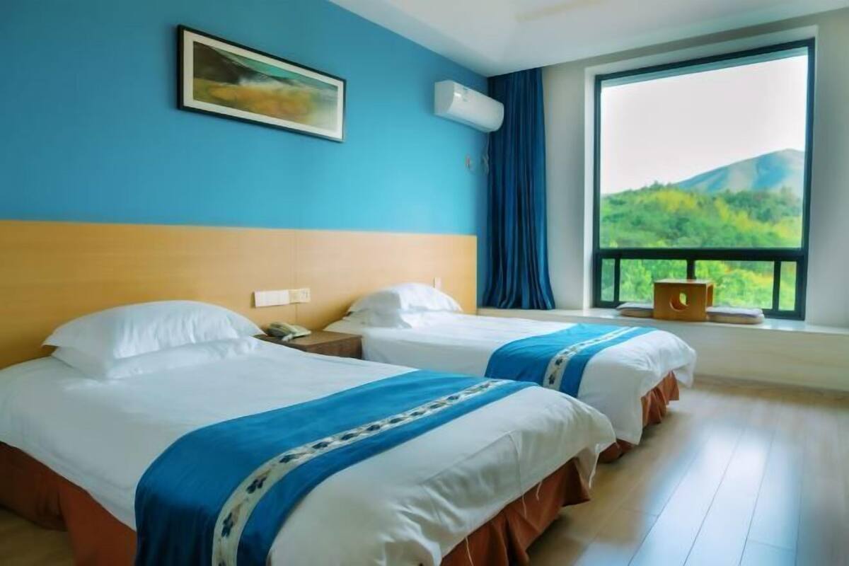 A hotel room with two beds, a painting on the wall, an air conditioning unit, a window with a view of greenery, and a wooden floor.