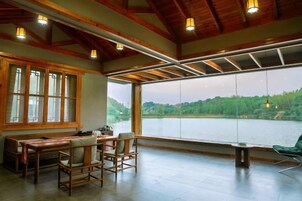 Amplia habitación con un gran ventanal con vistas a un lago, vigas de madera en el techo y una zona de comedor con sillas.