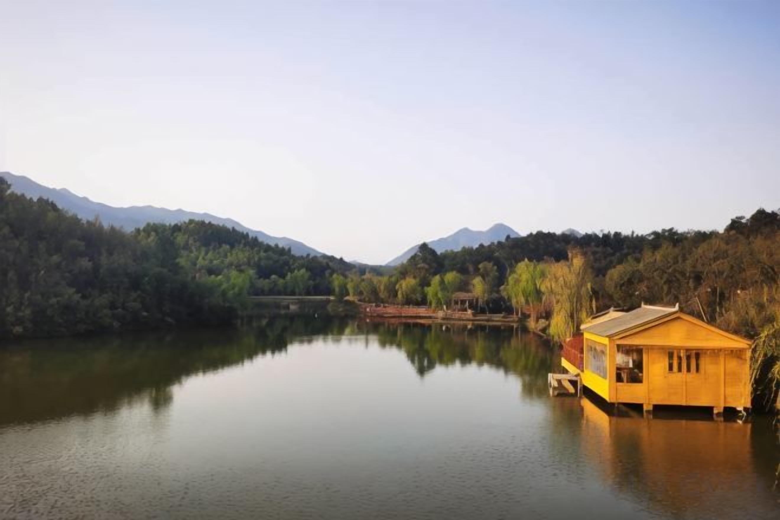 A tranquil lake with a wooden houseboat on the water, surrounded by forested hills.