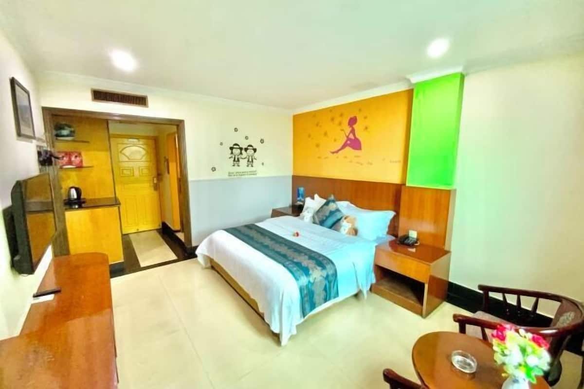 A hotel room with a bed, a desk, a chair, a small table with a vase, and a door leading to a bathroom.