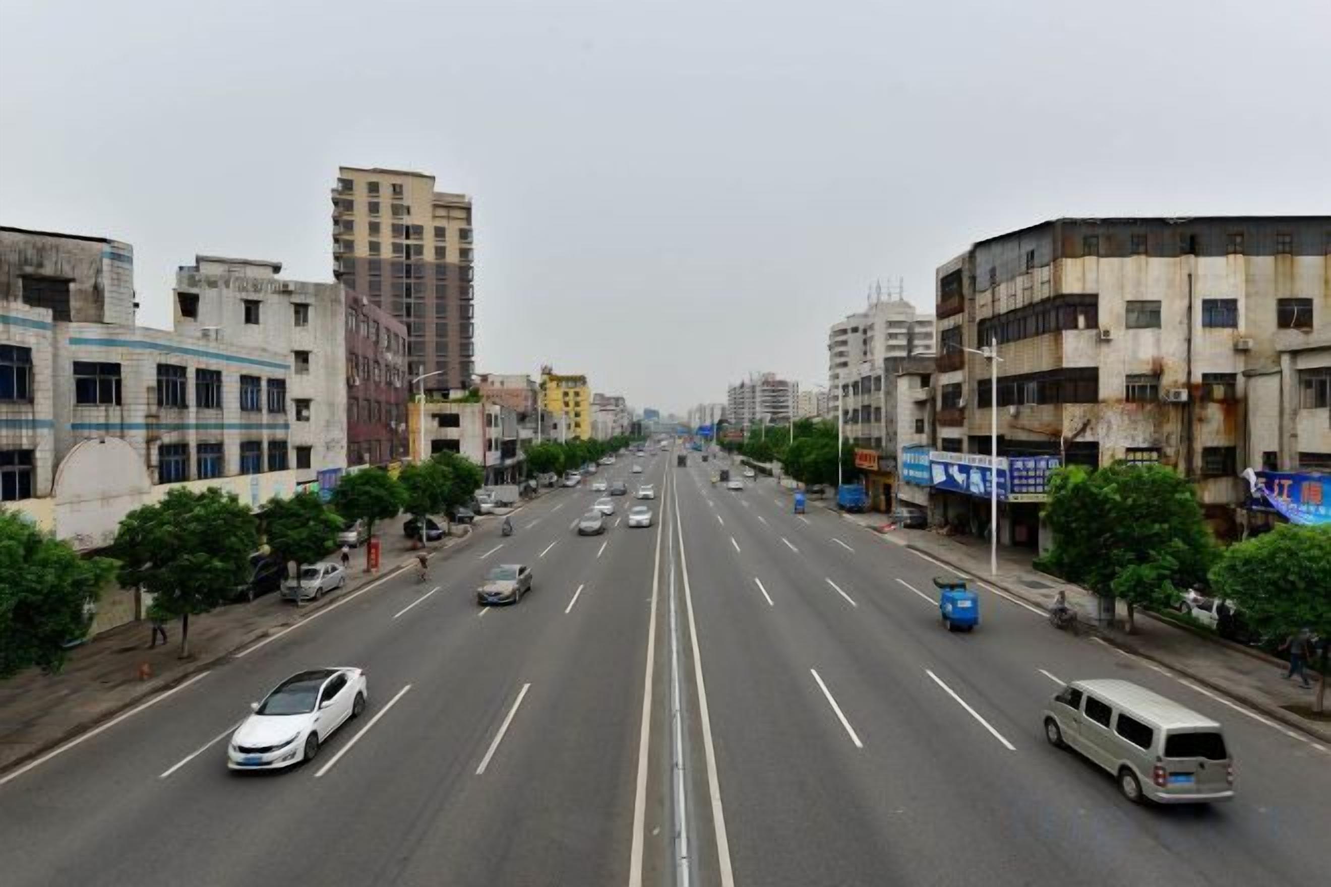A multi-lane urban road with vehicles, buildings, and trees.