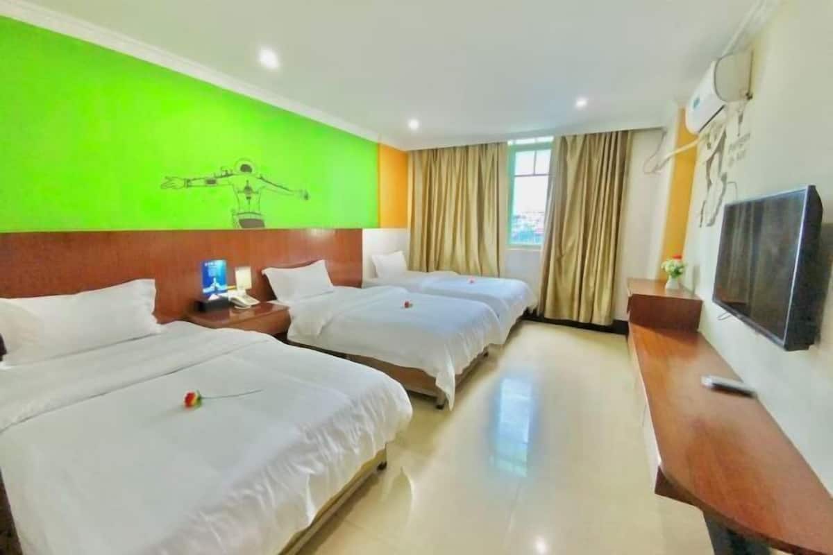 A hotel room with three beds, a wooden desk, a flat-screen TV, and a green wall with a drawing.