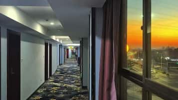 A hotel hallway with a carpeted floor and a large window with a city view at sunset.