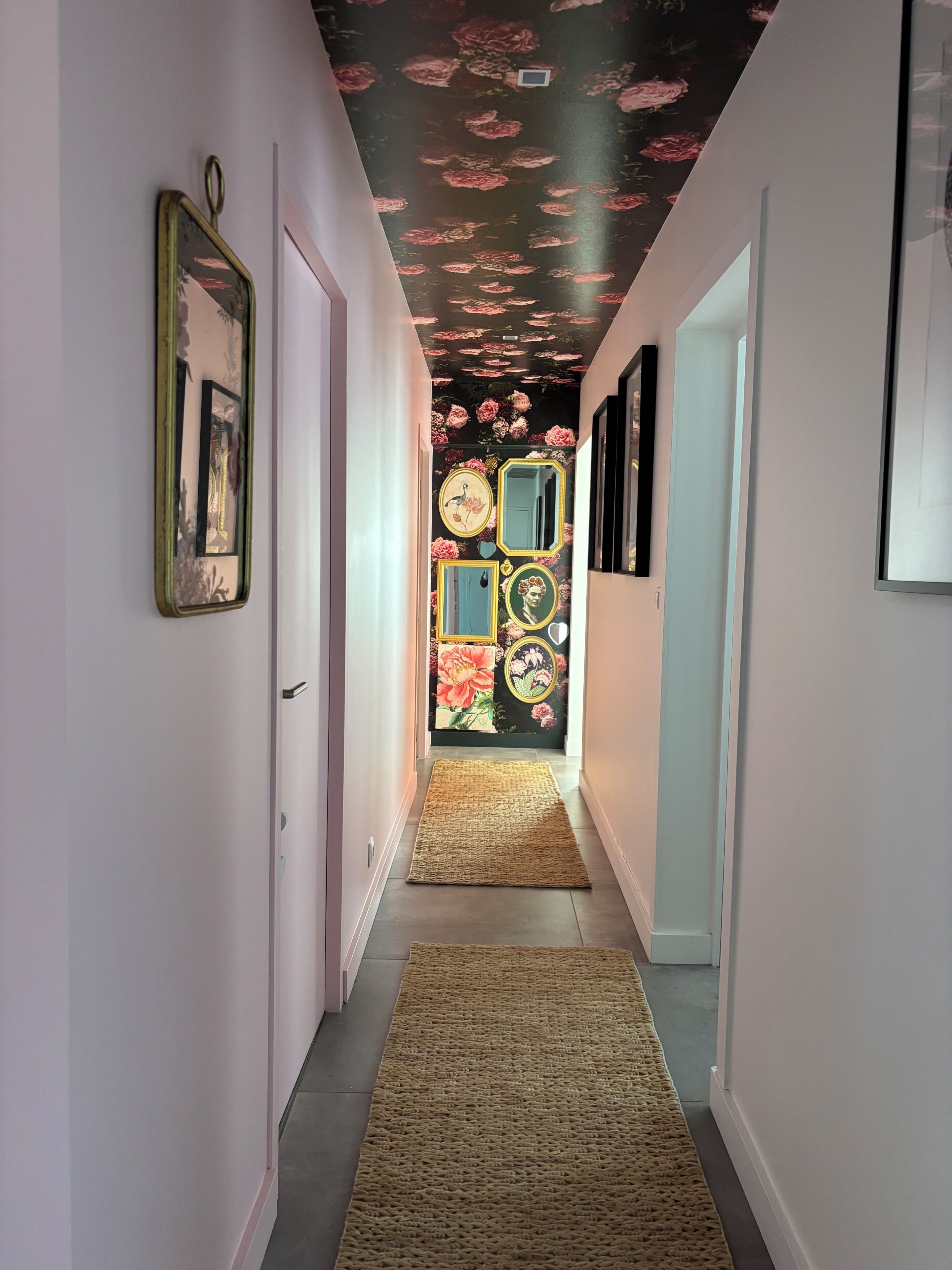 A hallway with a floral ceiling, framed pictures on the walls, and a woven rug on the floor.