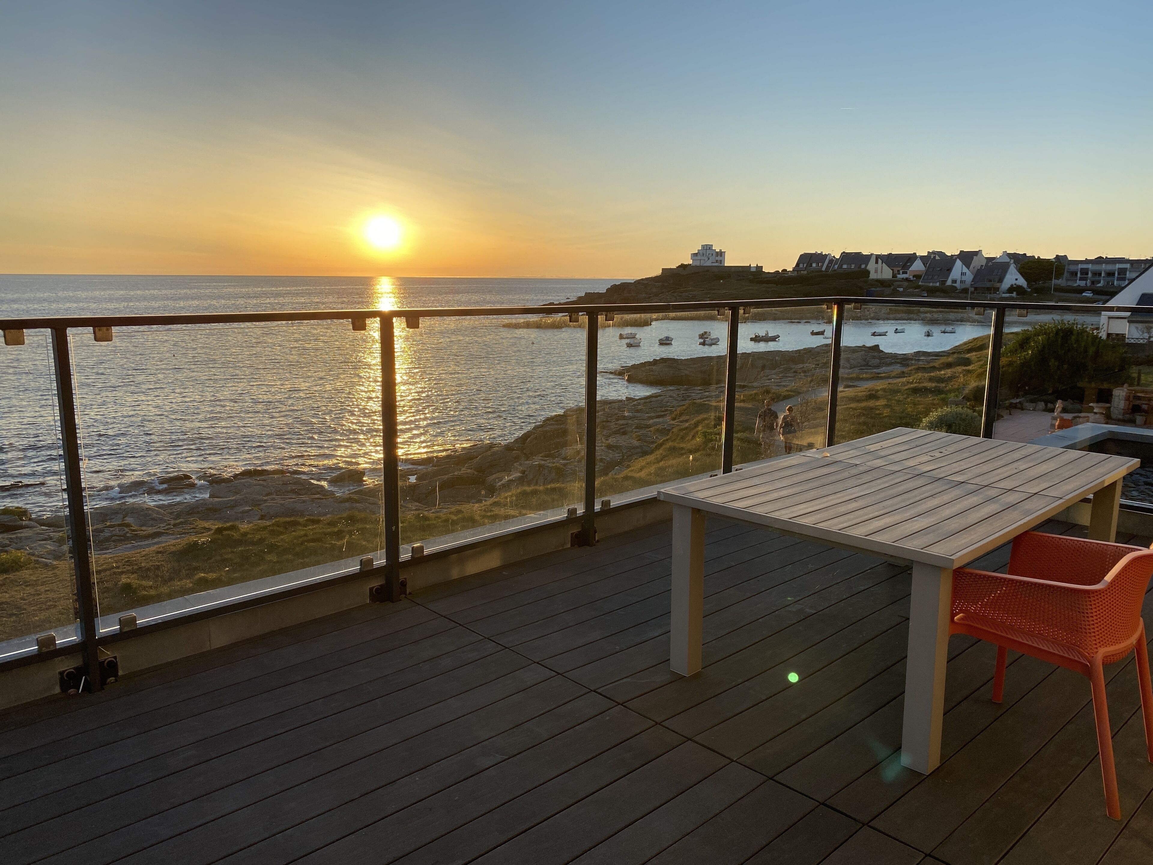 Eine Holzterrasse mit Tisch und Stühlen mit Blick auf eine Küstenlandschaft, im Hintergrund Boote und Gebäude.