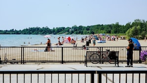 Una playa con gente nadando y tomando el sol, una valla en primer plano y una bicicleta estacionada cerca de la valla.