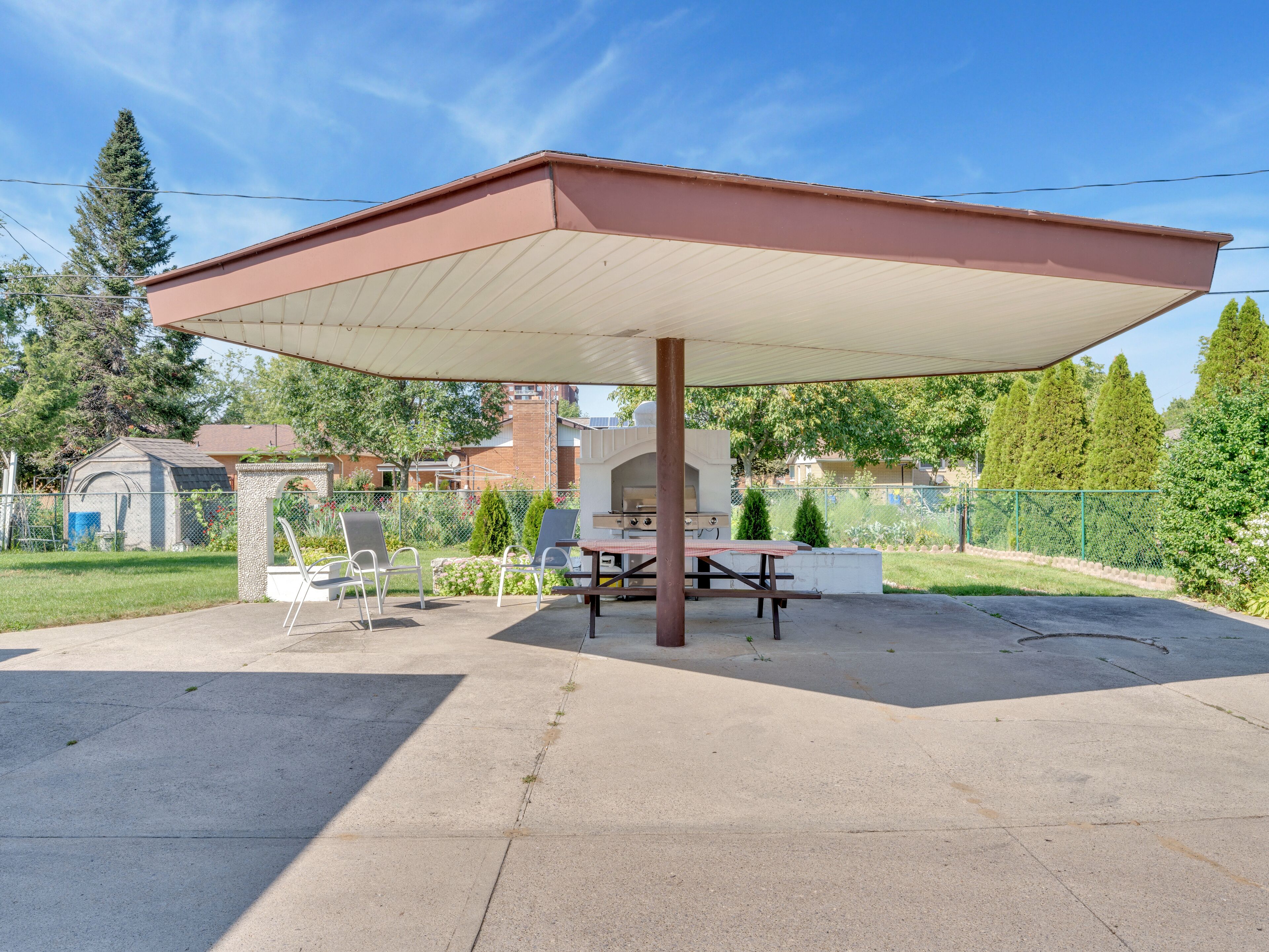 A covered outdoor area with a picnic table and chairs, surrounded by a fence and greenery.