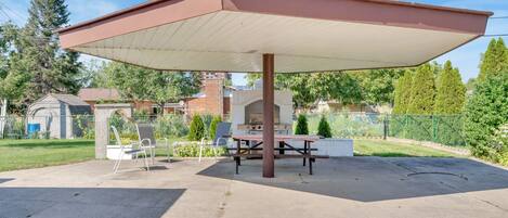 A covered outdoor area with a picnic table and chairs, surrounded by a fence and greenery.