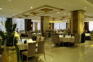Ampla sala de jantar com várias mesas e cadeiras, janelas grandes e plantas decorativas.
