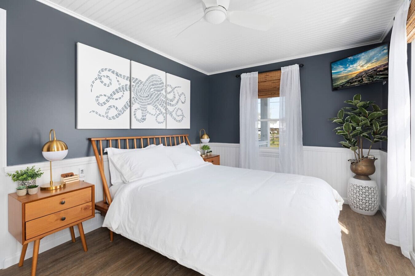 A bedroom with a bed, wooden nightstands, a ceiling fan, artwork, a window with curtains, and a potted plant.