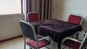 Una habitación con una mesa cubierta por un paño oscuro, cuatro sillas de metal con cojines rojos, una ventana con cortinas y una pared lisa.