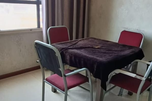 A room with a table covered by a dark cloth, four metal chairs with red cushions, a window with curtains, and a plain wall. - Weisi Haoting Hotel (Chibi)