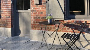 Une maison en briques avec un toit en tuiles, une porte en bois et une fenêtre. On peut voir à l’extérieur une petite table sur laquelle est posée une plante en pot, ainsi qu’une chaise pliante.