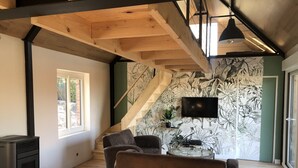 Un salon moderne avec un plafond en bois, un grand téléviseur fixé au mur et un escalier orné d’un papier peint à motifs floraux.