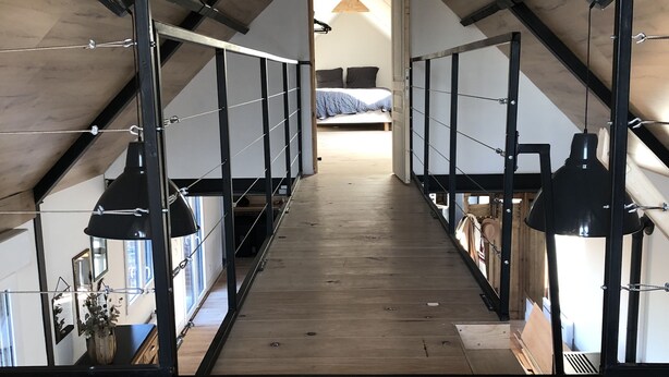 Une petite pièce mansardée au plancher de bois, avec un plafond en pente, des rampes de garde-corps en métal et un lit recouvert d’une couverture bleue.