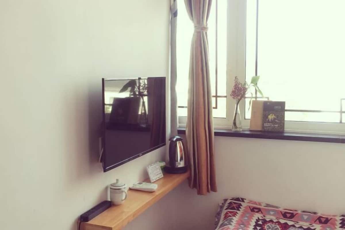 A room with a TV mounted on the wall, a shelf with a kettle, a small table with a phone and a cup, and a window with curtains.