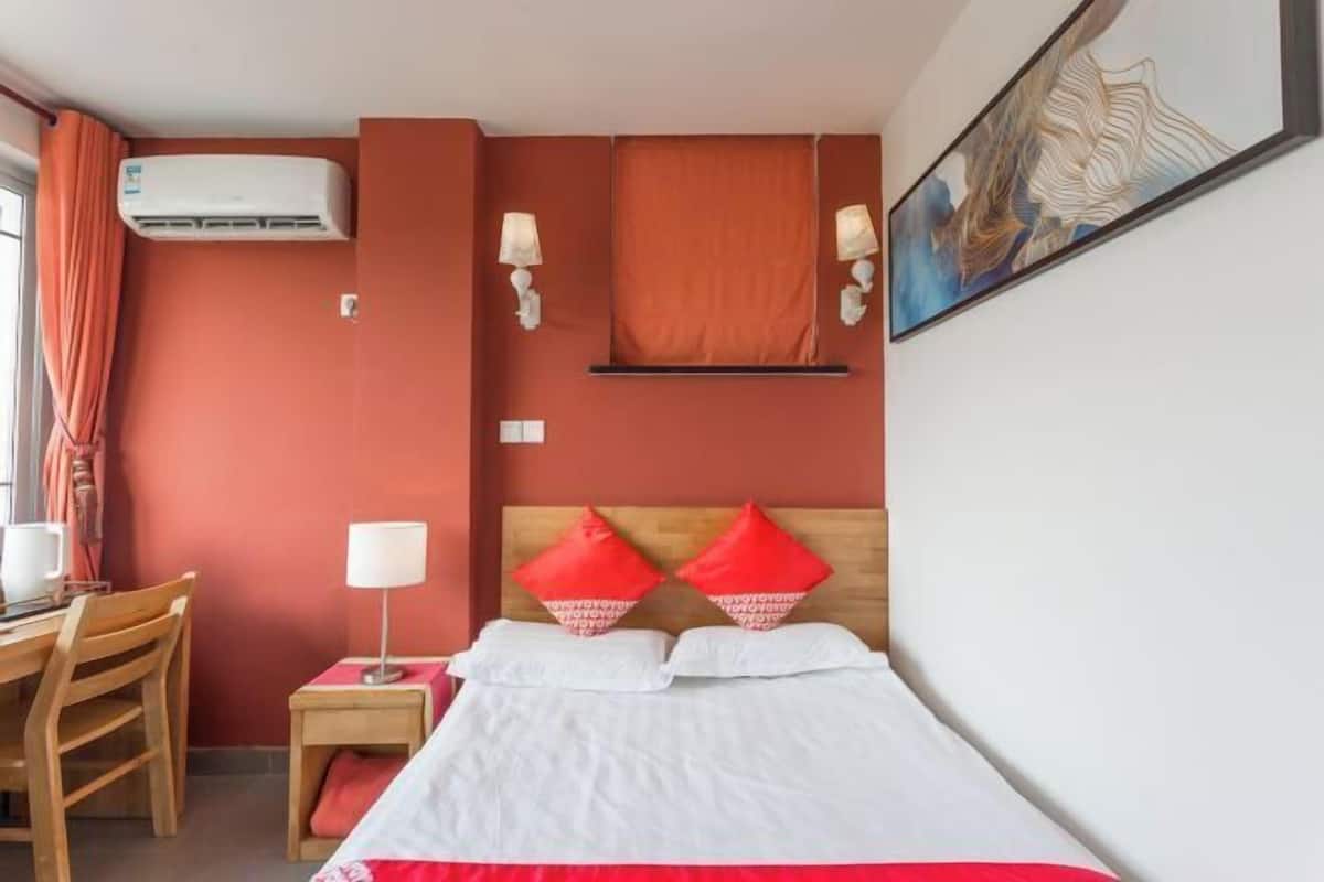 A hotel room with a bed, a desk, a chair, a nightstand, an air conditioner, and a painting on the wall.