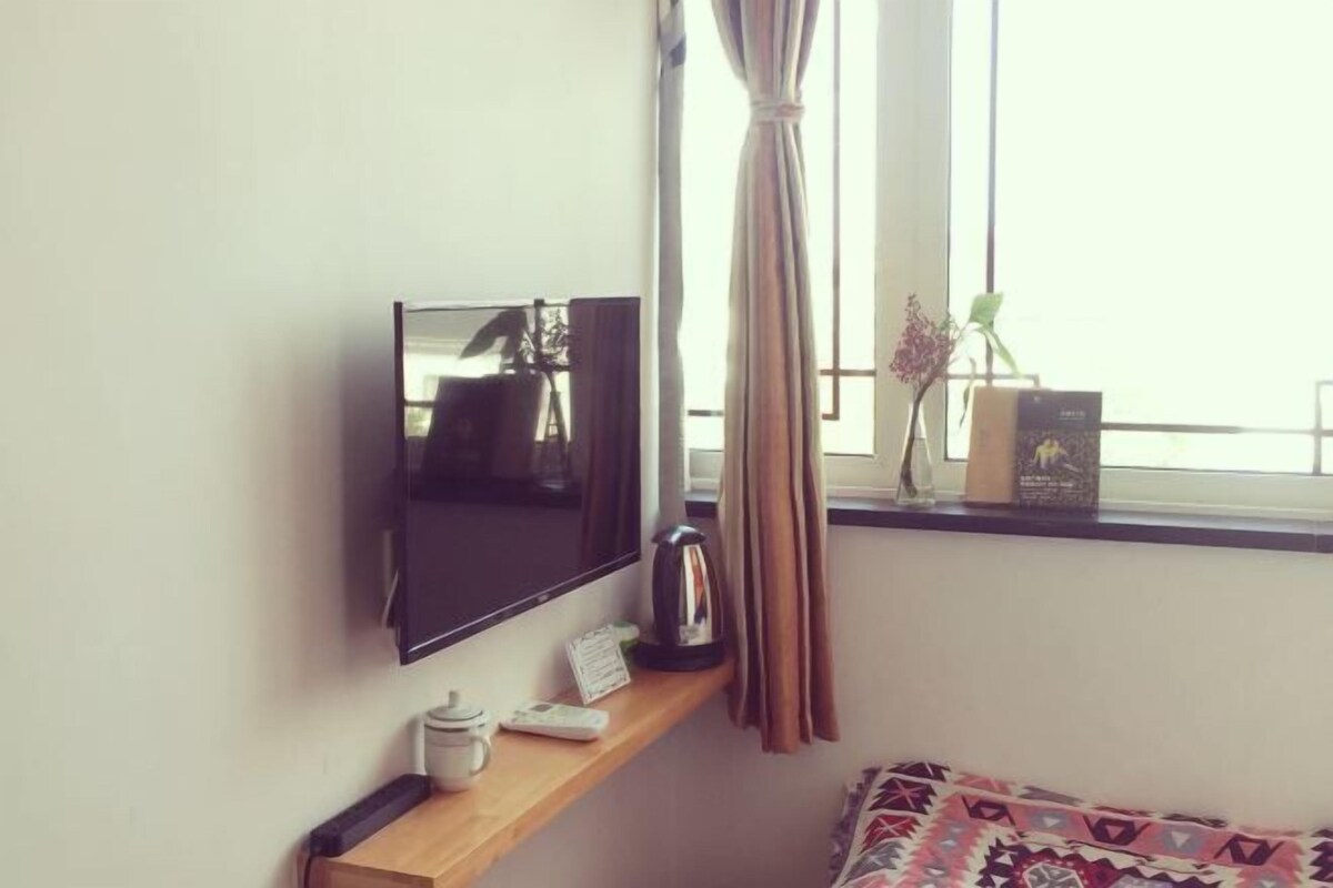 A room with a TV mounted on the wall, a shelf with a kettle, a small table, and a window with curtains.