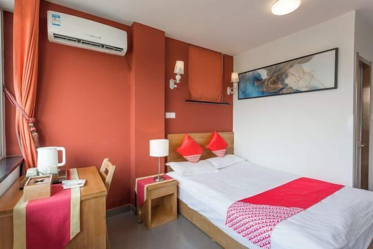 A hotel room with a bed, a desk, a chair, a nightstand, an air conditioning unit, and a painting on the wall.