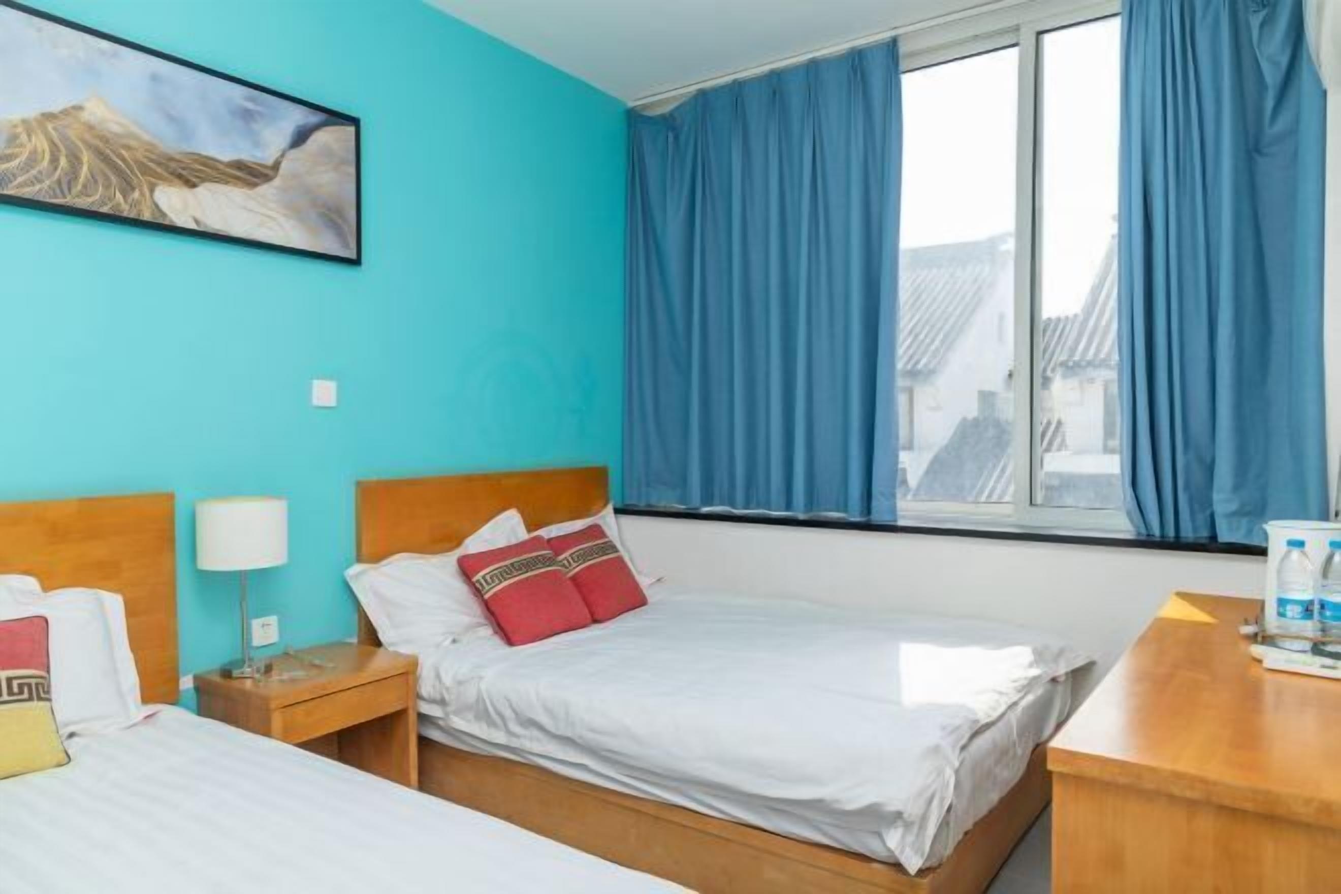 A hotel room with two beds, a window with curtains, a wooden headboard, a bedside table with a lamp, and a painting on the wall.