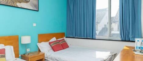 A hotel room with two beds, a window with curtains, a wooden headboard, a bedside table with a lamp, and a painting on the wall.