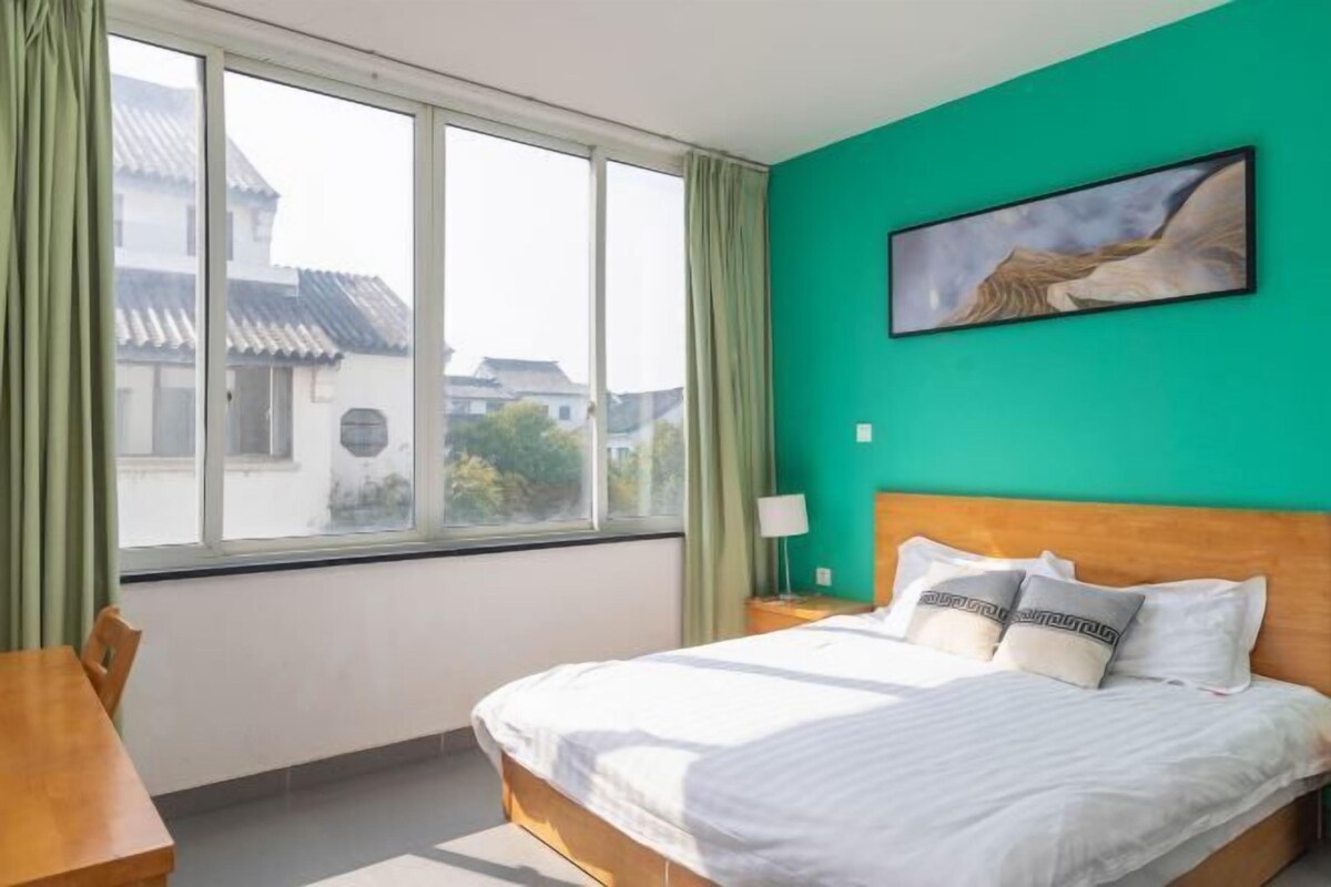 A hotel room with a large window, a bed with white bedding, a green wall, and a painting above the bed.