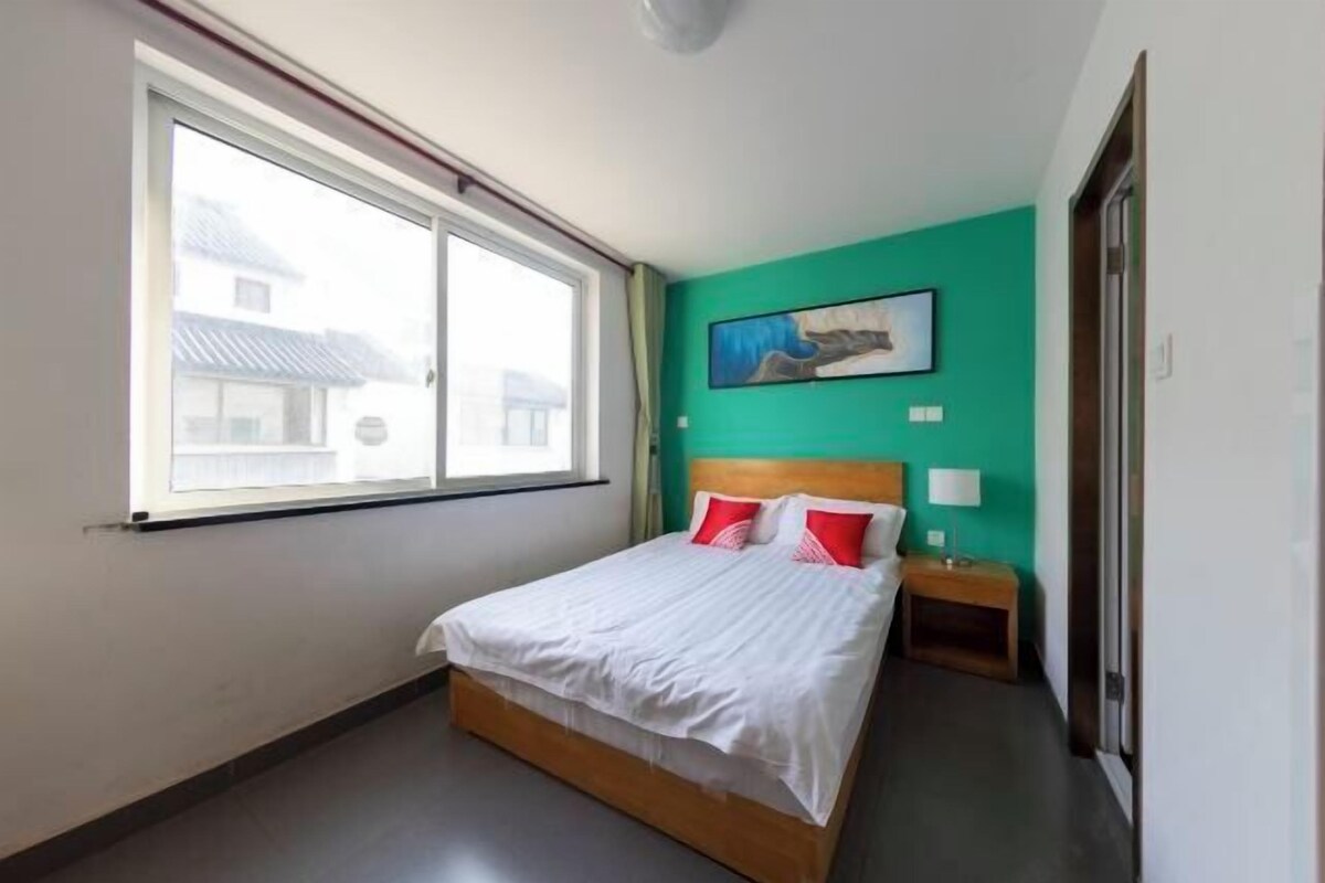 A bedroom with a large window, a bed with white bedding and red pillows, a wooden headboard, a nightstand with a lamp, and a painting on the teal wall.