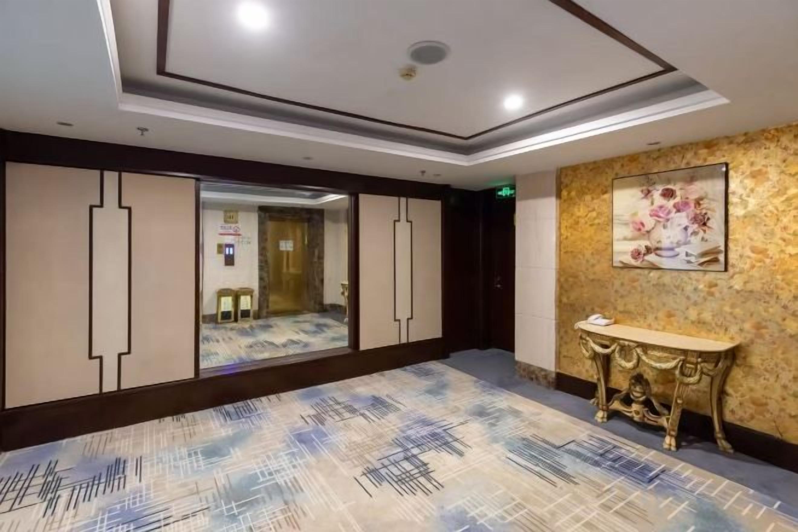 A hotel lobby with a patterned carpet, a reception desk, a framed artwork on the wall, and a door leading to another room.