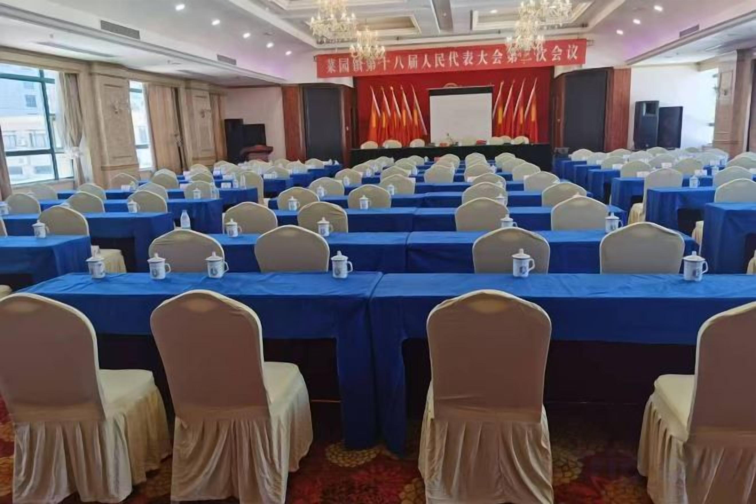 Un salón de conferencias con filas de mesas y sillas, un escenario con un telón de fondo rojo y un cartel.