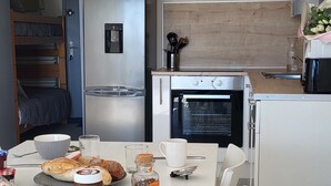 Cocina compacta con refrigerador, horno y microondas. Hay una mesa de comedor con productos para el desayuno y un reloj en la pared.