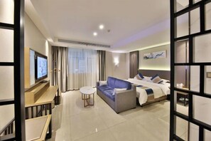 A hotel room with a bed, a sofa, a TV, a small table, and a window with curtains.