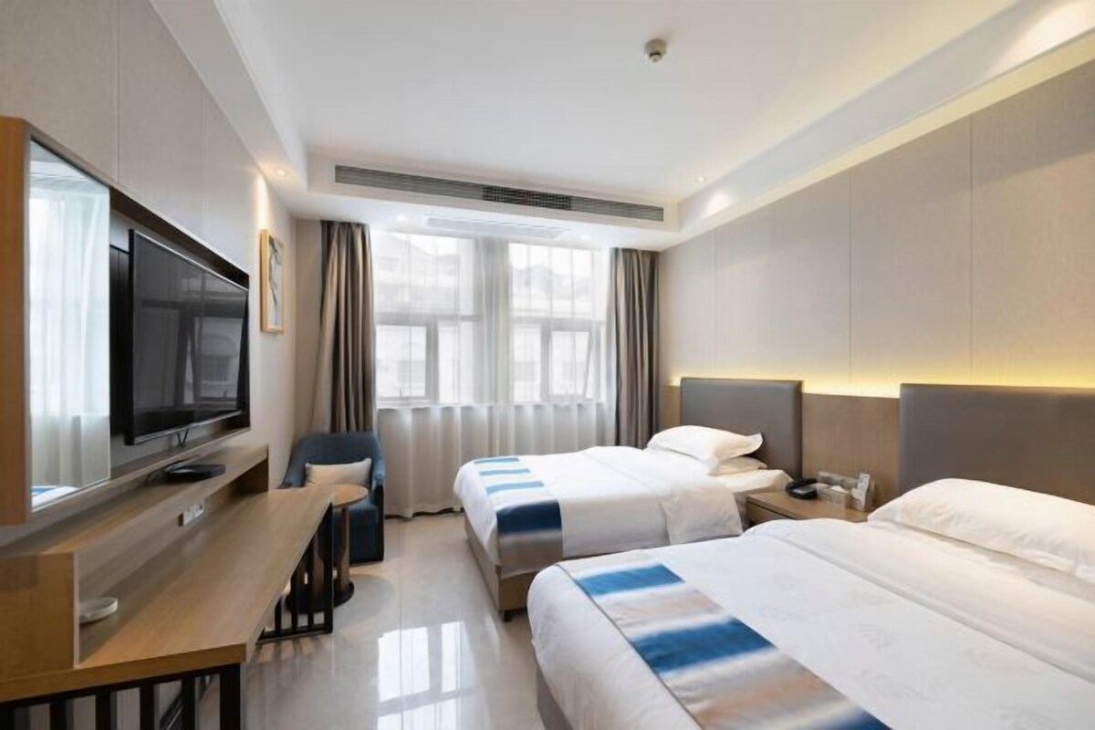 A hotel room with two beds, a TV, a desk, and a large window with curtains.