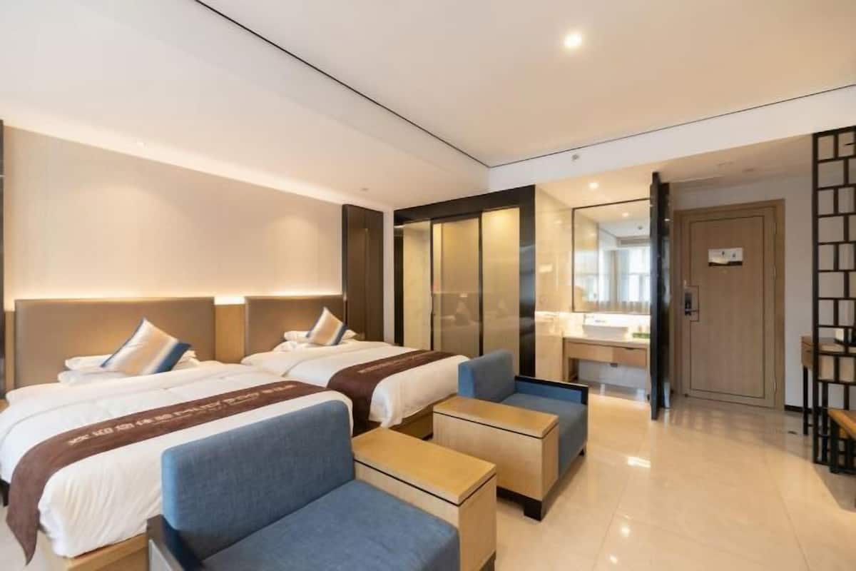 Une chambre d’hôtel moderne avec deux lits, un coin salon comprenant un canapé et une petite table, une salle de bain avec un miroir et un lavabo, et une porte donnant sur une autre pièce.